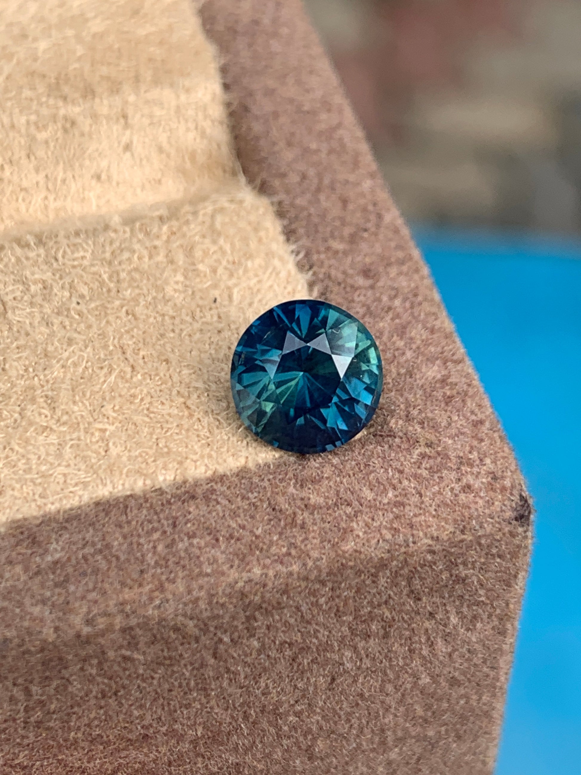 Un saphir sarcelle rond de 2,4 carats de Madagascar de Bonnot Paris repose sur une surface brune texturée, mettant en valeur son bleu profond avec des reflets verts sur un fond bleu flou.