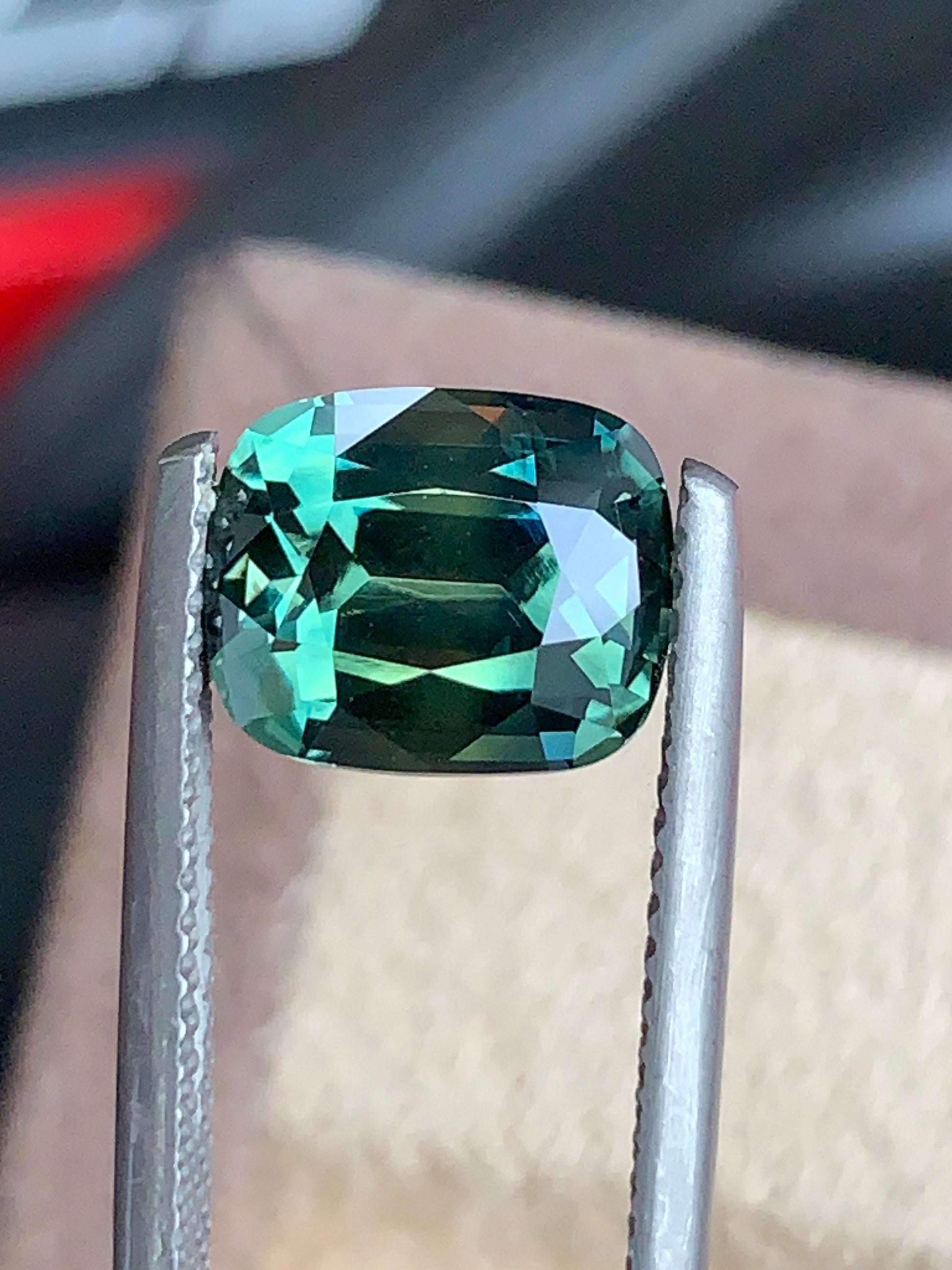 Gros plan sur la pierre précieuse « Saphir Teal • 4,2 ct » de Bonnot Paris, une pierre précieuse éclatante taille coussin, maintenue par une pince métallique. Ce trésor non chauffé de Madagascar met en valeur ses facettes sur un fond neutre et flou.