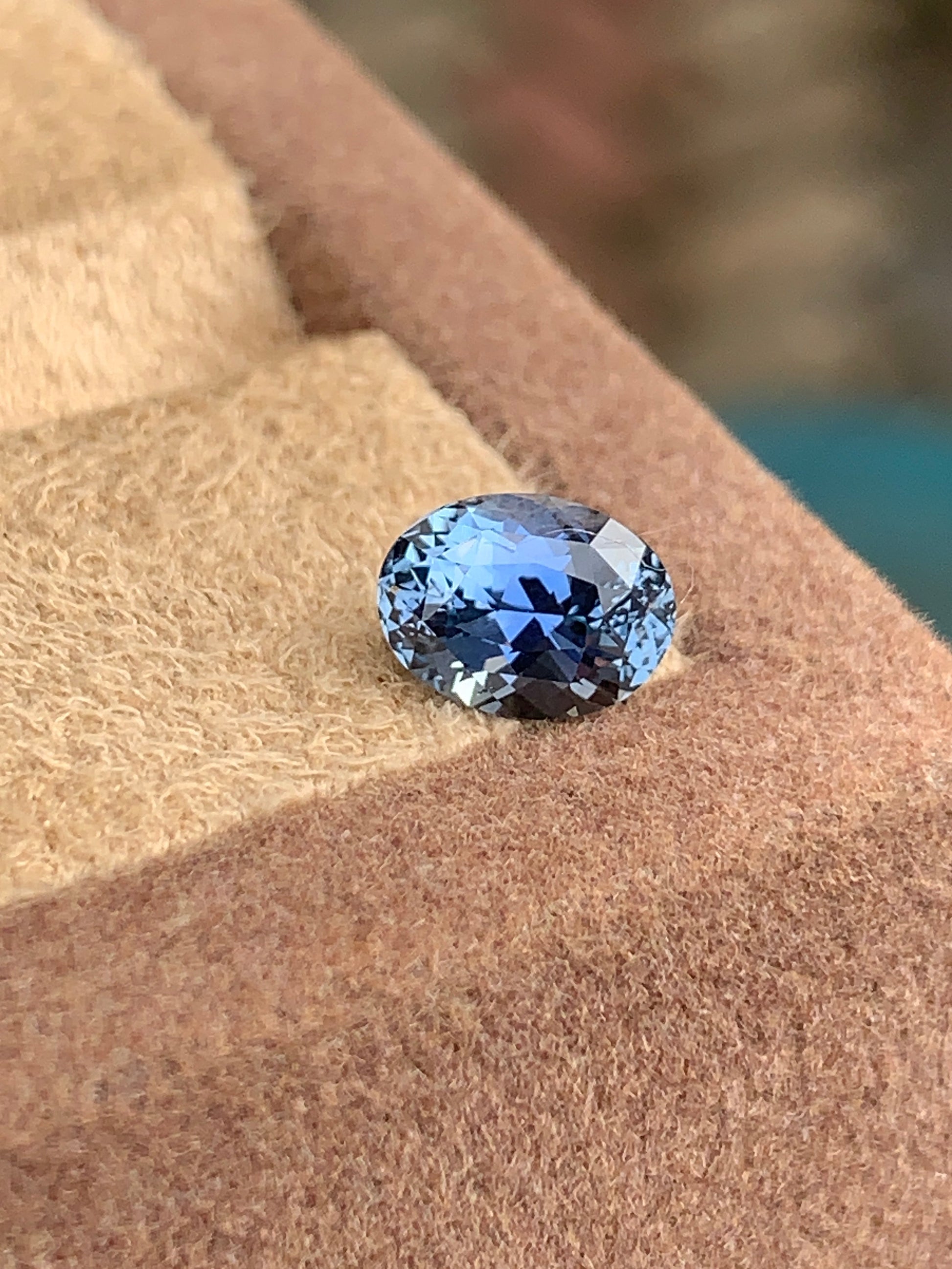 Le Saphir Bleu • 1,7ct de Bonnot Paris, une pierre précieuse bleue ovale à facettes, repose sur une surface texturée beige et marron, reflétant la lumière.