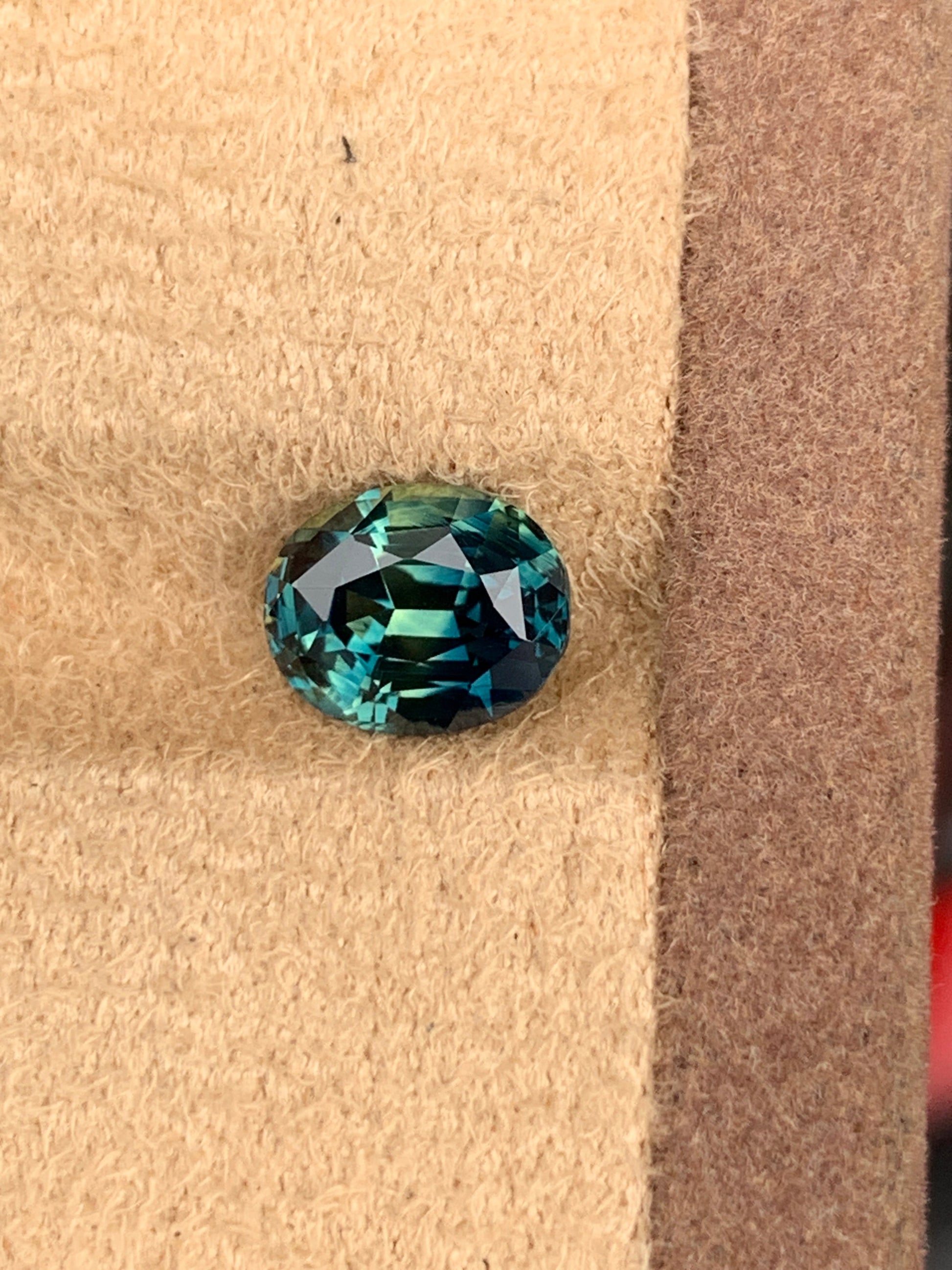 La pierre précieuse Saphir Teal de Bonnot Paris, de 2,7 carats, présente un design rond et facetté sur une surface beige texturée et une couleur vibrante et profonde. Ses tailles complexes reflètent brillamment la lumière, créant un éclat enchanteur.