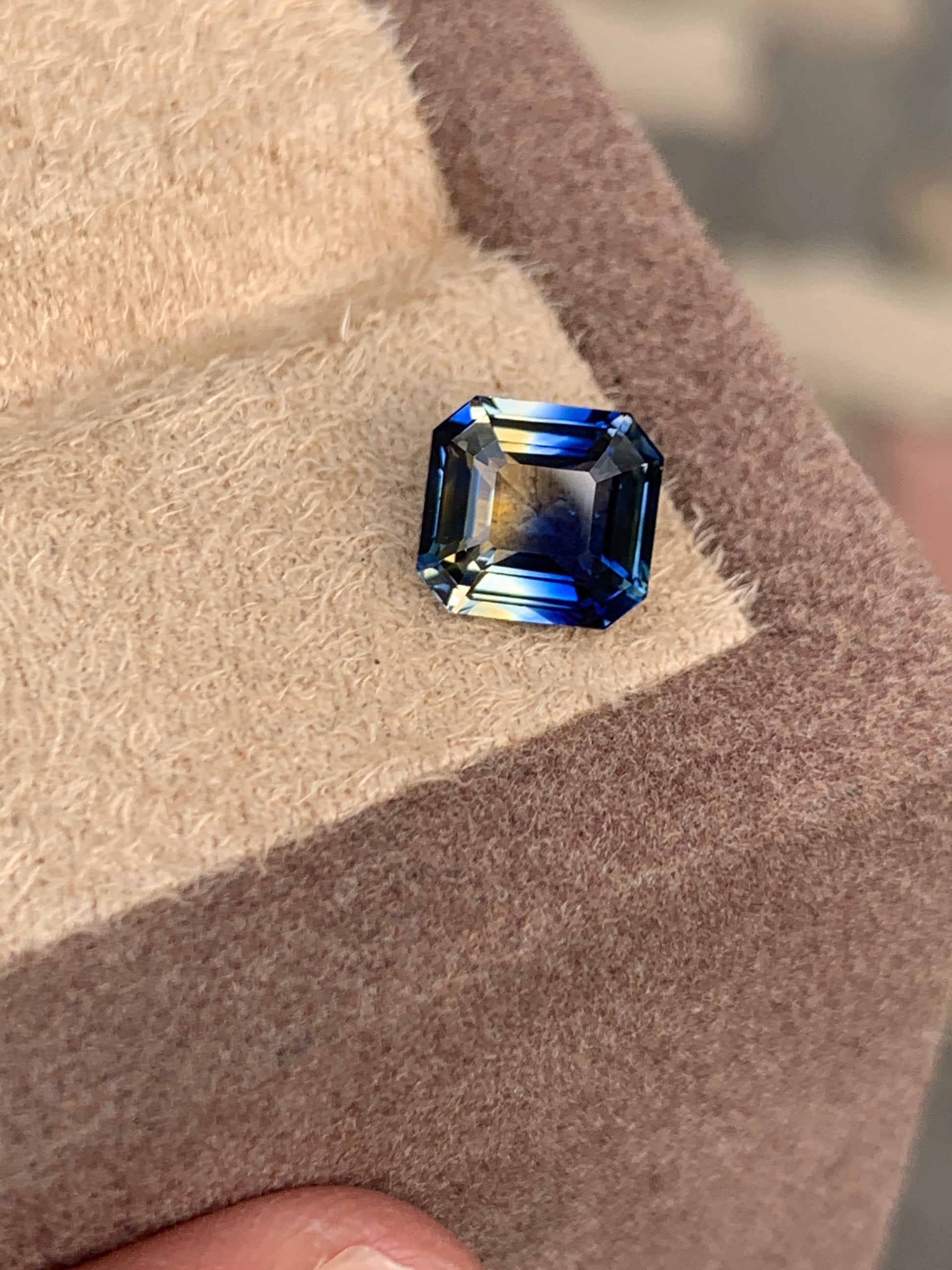 Un Saphir bicolore rectangulaire taille émeraude de 3,01 ct de Bonnot Paris, avec des touches de bleu et de vert, repose en biais sur une surface de velours beige sur un fond légèrement flou.