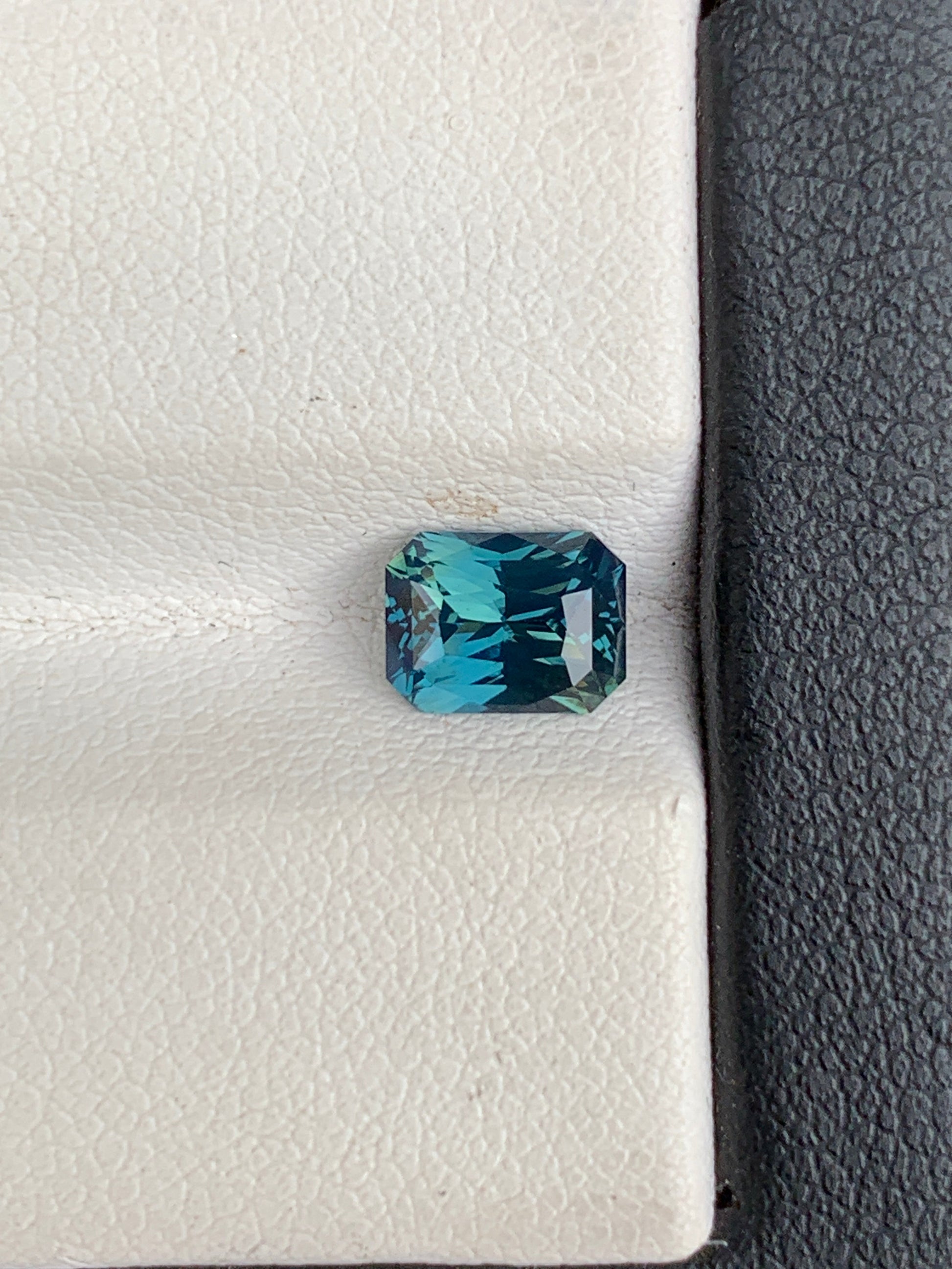 Le Saphir Teal de 1,52 ct de Bonnot Paris, avec sa taille émeraude bleu-vert vibrante, brille sur un fond noir et blanc texturé, mettant en valeur ses facettes complexes et sa riche profondeur de couleur.