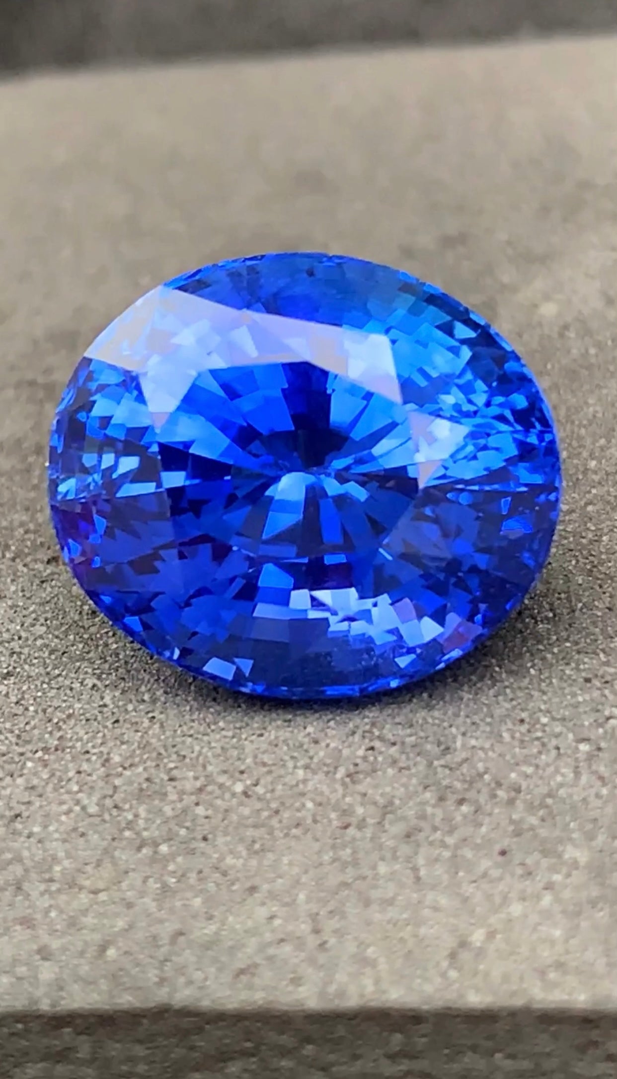 Le "Saphir Bleu • 14,1ct" de Bonnot Paris est une pierre précieuse facettée d'un bleu profond, de forme ovale, reposant sur une surface grise, reflétant la lumière avec des facettes brillantes qui mettent en valeur sa teinte vive et sa clarté.