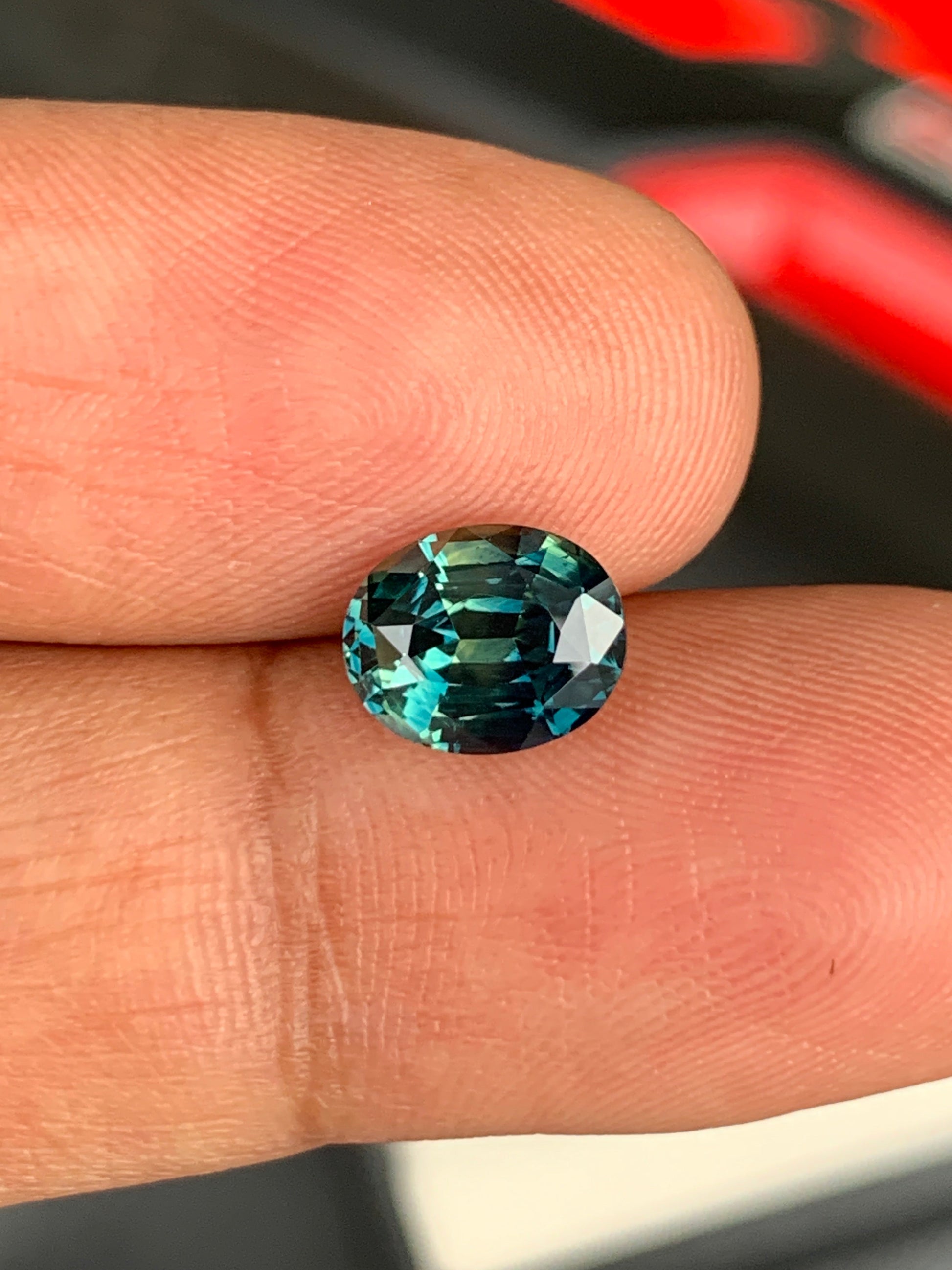 Une main tient une pierre précieuse Saphir Teal de 2,7 carats de Bonnot Paris, dont la taille ovale multifacette scintille de différentes nuances de bleu. Le flou d'arrière-plan souligne l'éclat de la pierre.