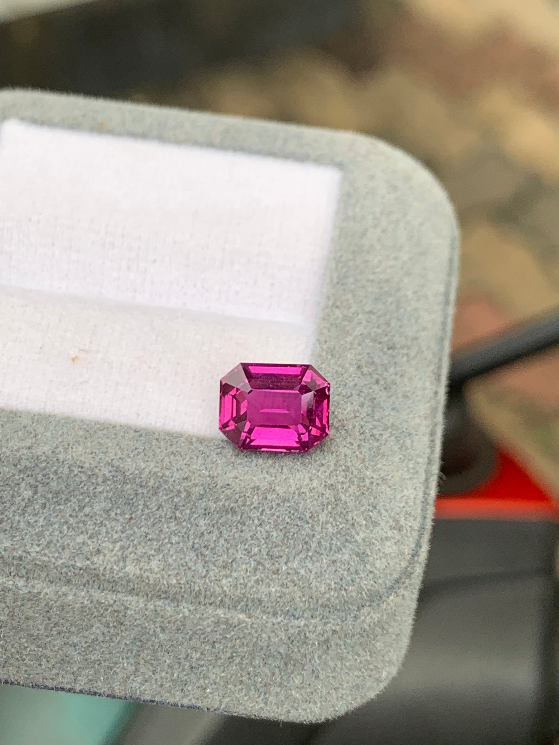Un Saphir rose éclatant de 3,01 ct de Bonnot Paris, taille émeraude, repose sur un coussin gris doux dans un écrin ouvert. Le fond légèrement flou met en valeur la couleur riche et la surface facettée de la pierre précieuse.