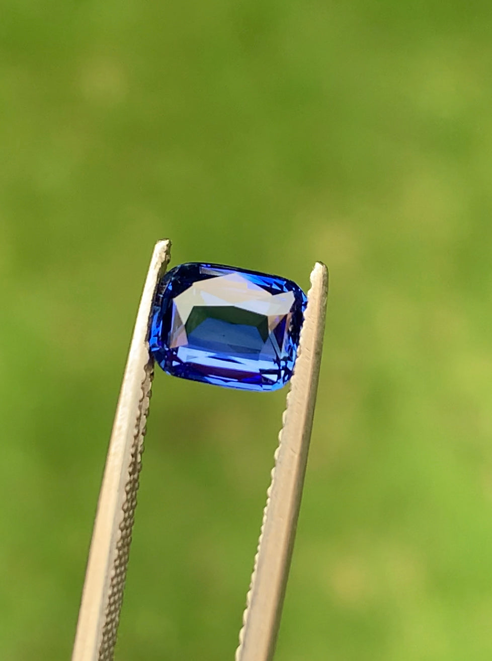 Le Saphir Bleu • 1,57 ct de Bonnot Paris est maintenu par une pincette sur un fond vert flou. La pierre rectangulaire aux angles biseautés présente des facettes reflétant la lumière dans des nuances de bleu.