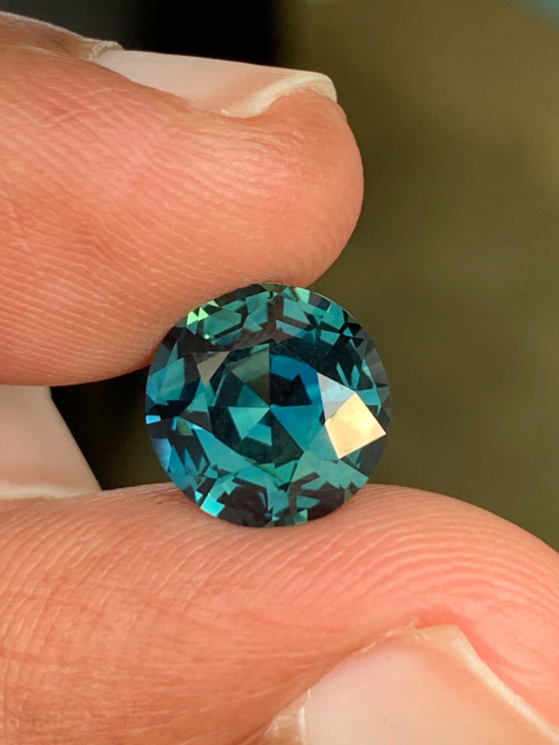 Un gros plan capture une main tenant l'éblouissant Saphir Teal de 4,1 carats de Bonnot Paris, une gemme de Madagascar non chauffée aux reflets bleus et verts profonds. Ses tailles complexes et sa clarté reflètent magnifiquement la lumière, mettant en valeur son éclat.