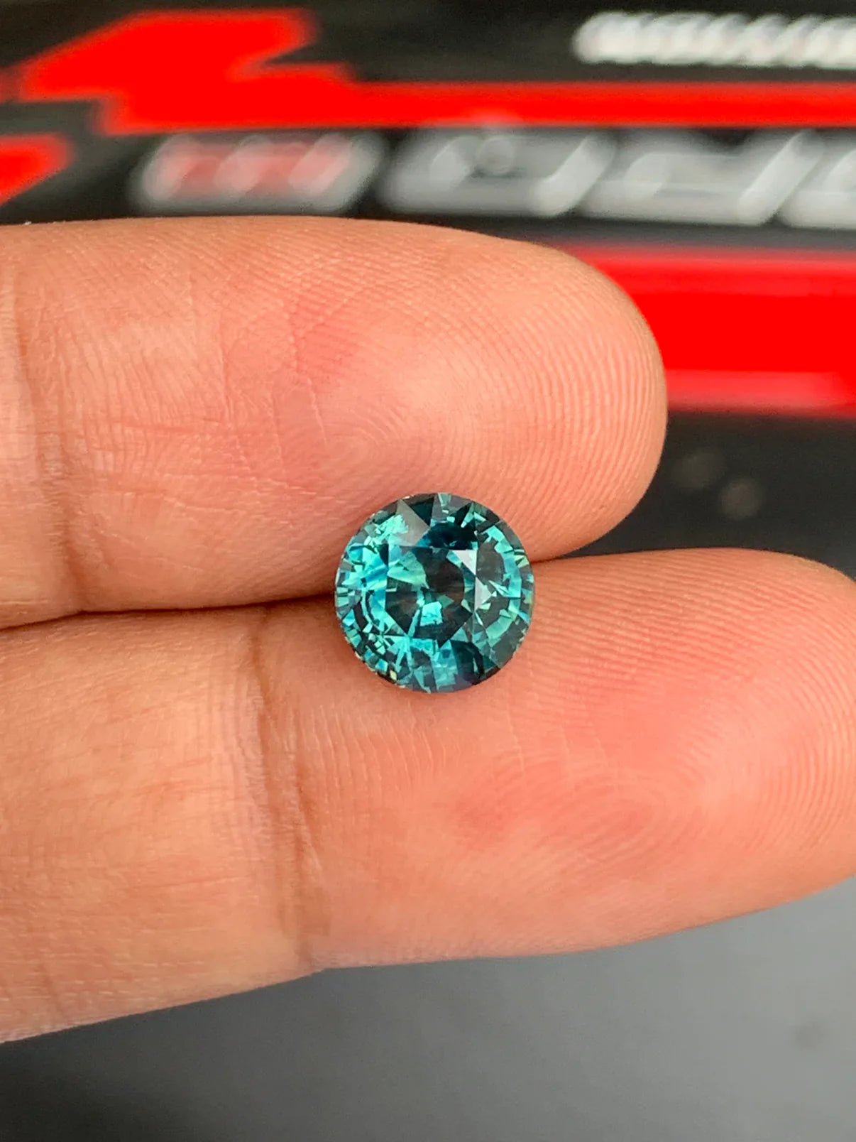 Une personne tient entre le pouce et l'index le saphir turquoise de Madagascar de 3,6 carats de Bonnot Paris. La pierre ronde, taille brillant, scintille sous la lumière, révélant des facettes complexes sur un fond flou de nuances rouges et noires.