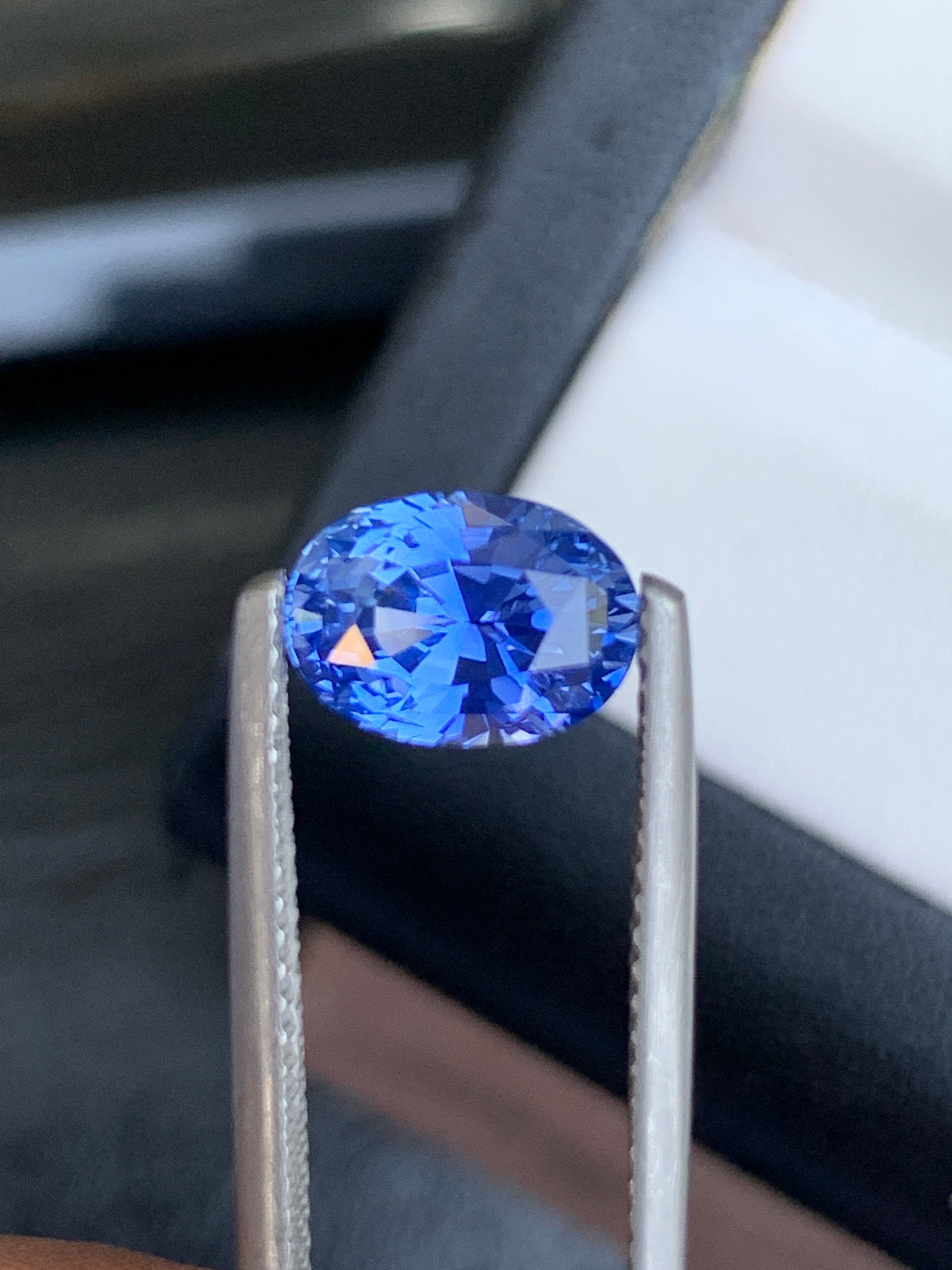 Une image montre une pince à épiler tenant un Saphir Bleu étincelant de 3,02 carats de Bonnot Paris, un bijou ovale sur mesure aux superbes nuances de bleu. Ses facettes scintillent à la lumière, tandis qu'un sertissage sombre révèle un écrin partiellement visible, ajoutant à l'intrigue.