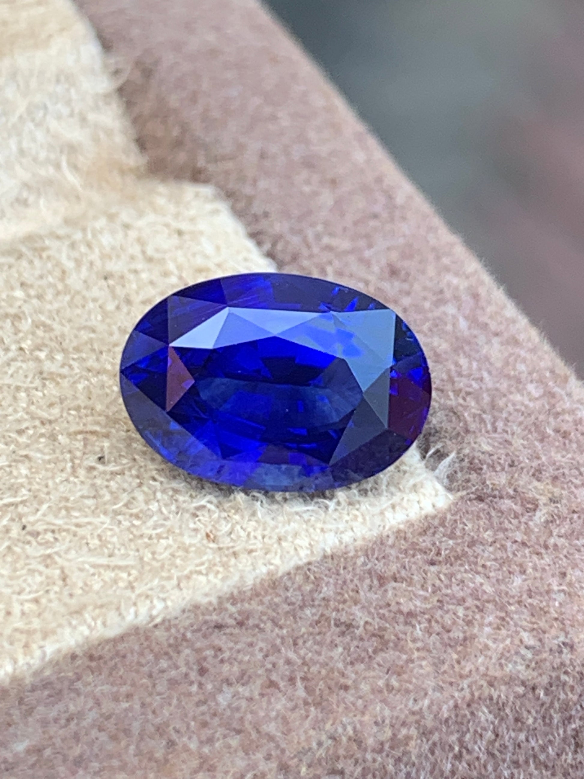 Gros plan sur le Saphir Bleu • 3,51ct taille ovale de Bonnot Paris qui scintille d'un bleu profond sur une surface texturée beige, sur fond brun flou.