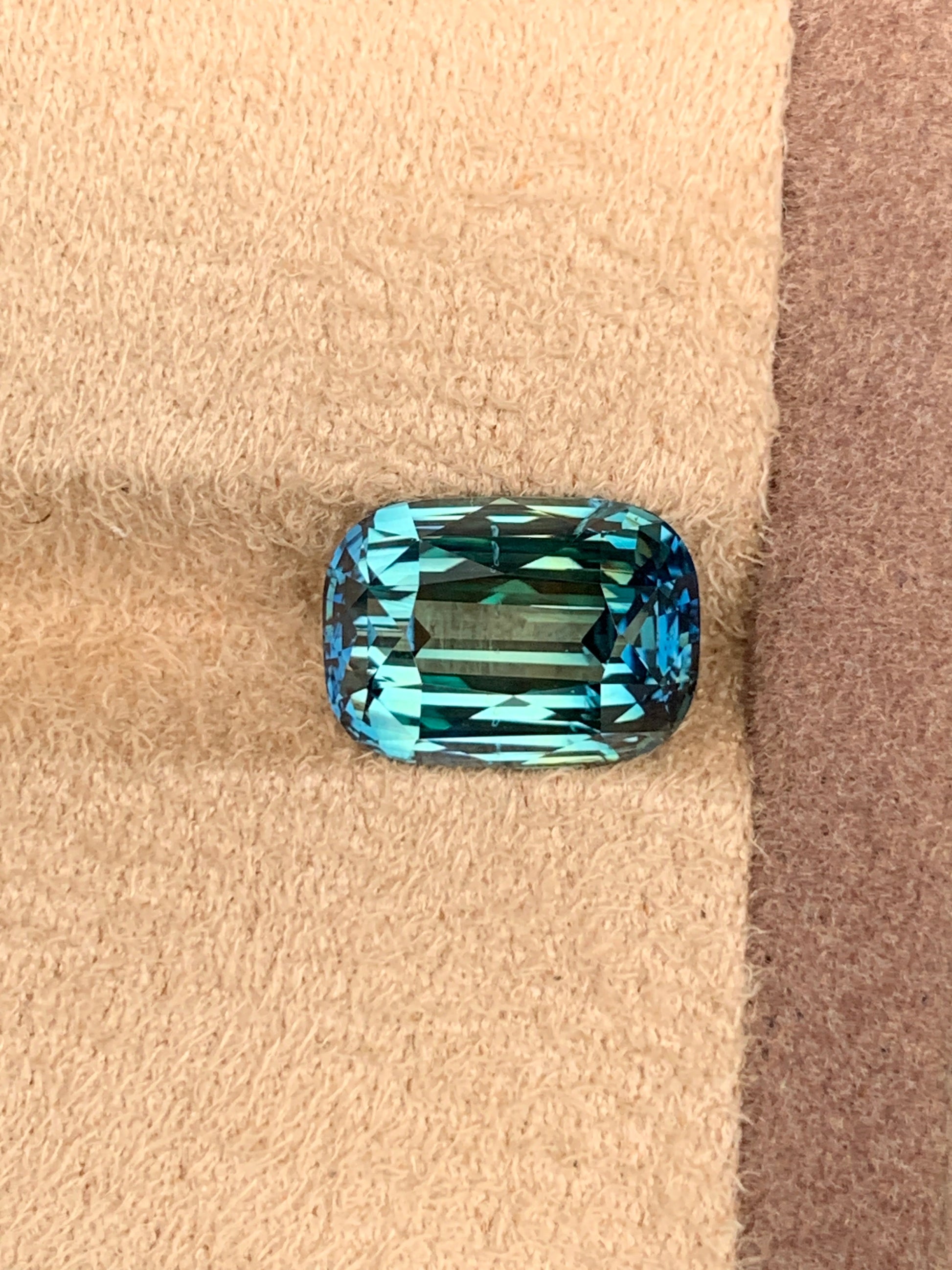 La pierre précieuse Saphir Teal • 7,6 ct de Bonnot Paris est un bijou rectangulaire facetté de couleur bleu saphir profond sarcelle, posé sur une surface texturée marron clair et captant la lumière pour révéler sa clarté et ses coupes complexes.