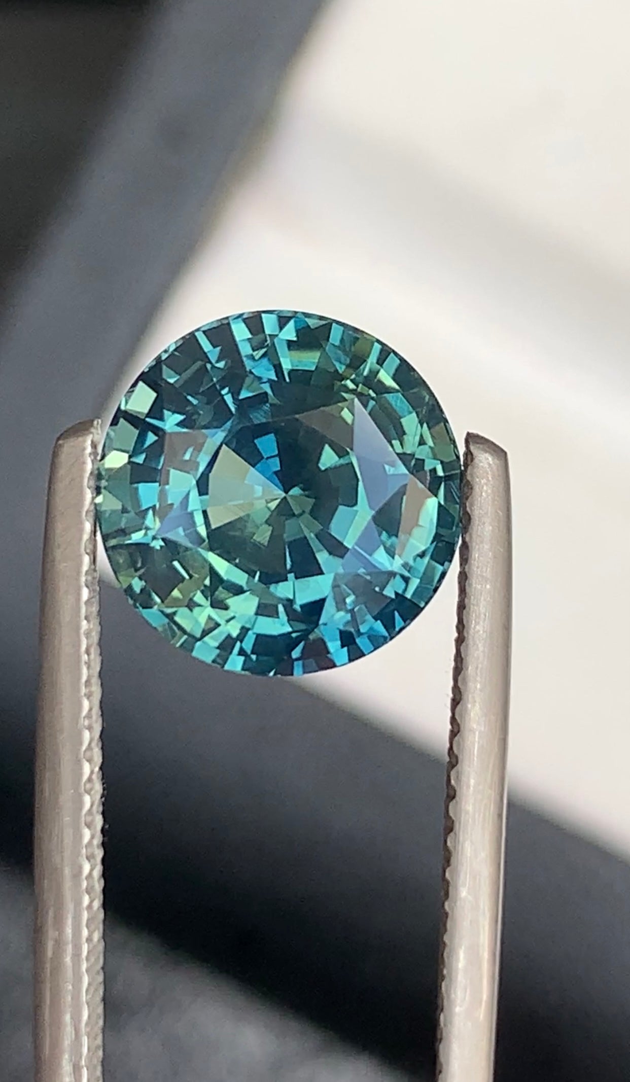 Une paire de pinces en métal maintient l'exquise pierre précieuse Bonnot Paris Saphir Teal • 5,52 ct sur un fond flou, où elle scintille dans des tons de turquoise et de sarcelle, soulignant sa taille complexe et sa clarté.