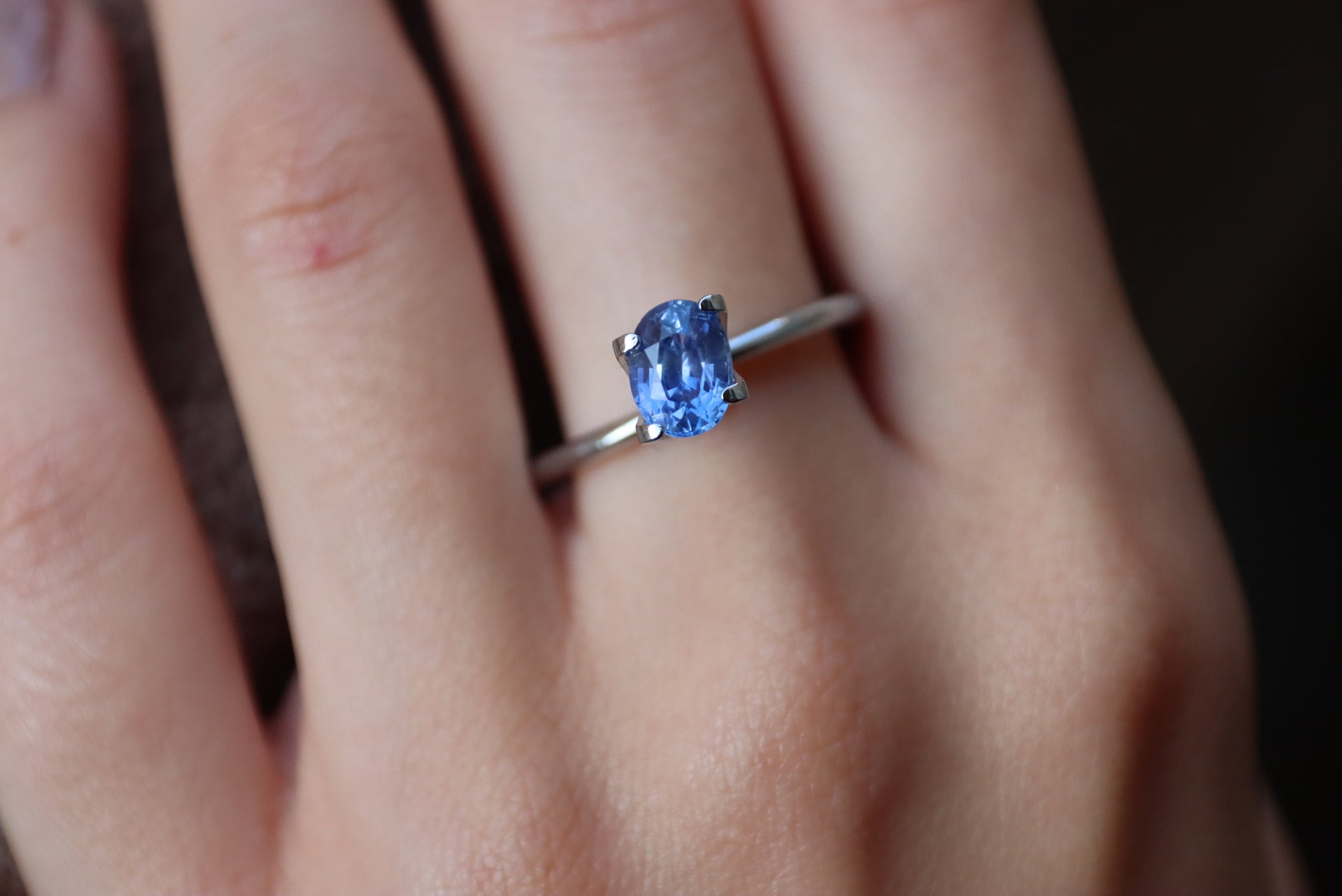 Une main ornée de la bague Saphir Bleu · 1,28 ct de Bonnot Paris, comportant une pierre précieuse bleue rectangulaire taillée en brillant et sertie en argent, repose gracieusement sur l'annulaire sur un fond sombre et flou.