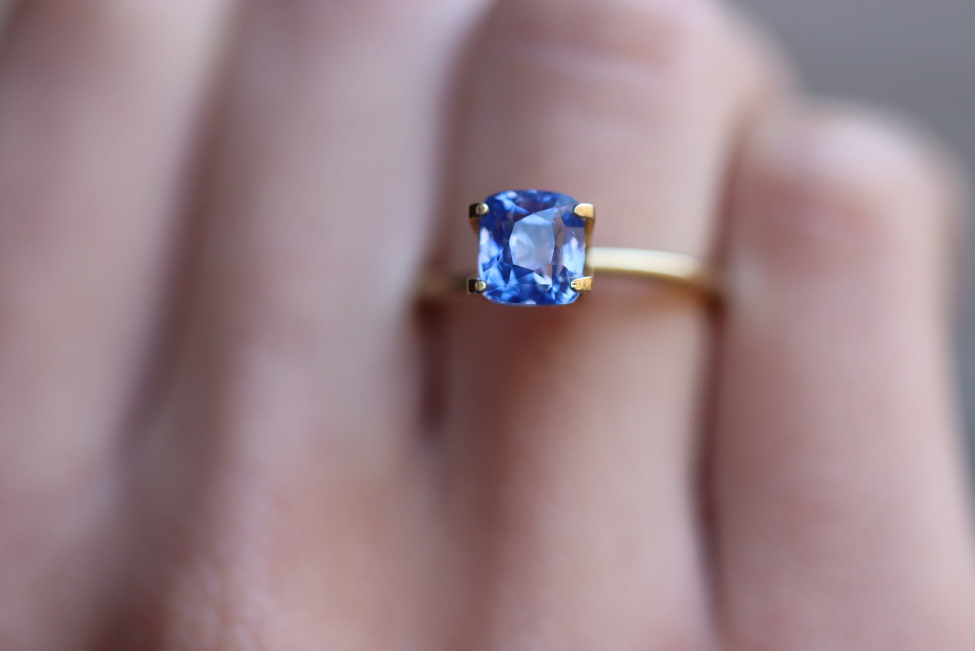 La bague Saphir Bleu · 3,03 ct de Bonnot Paris, une bague en or avec une pierre précieuse bleue taille coussin dans un sertissage à quatre griffes, est présentée sur un doigt avec l'arrière-plan légèrement flou pour accentuer la gemme scintillante.