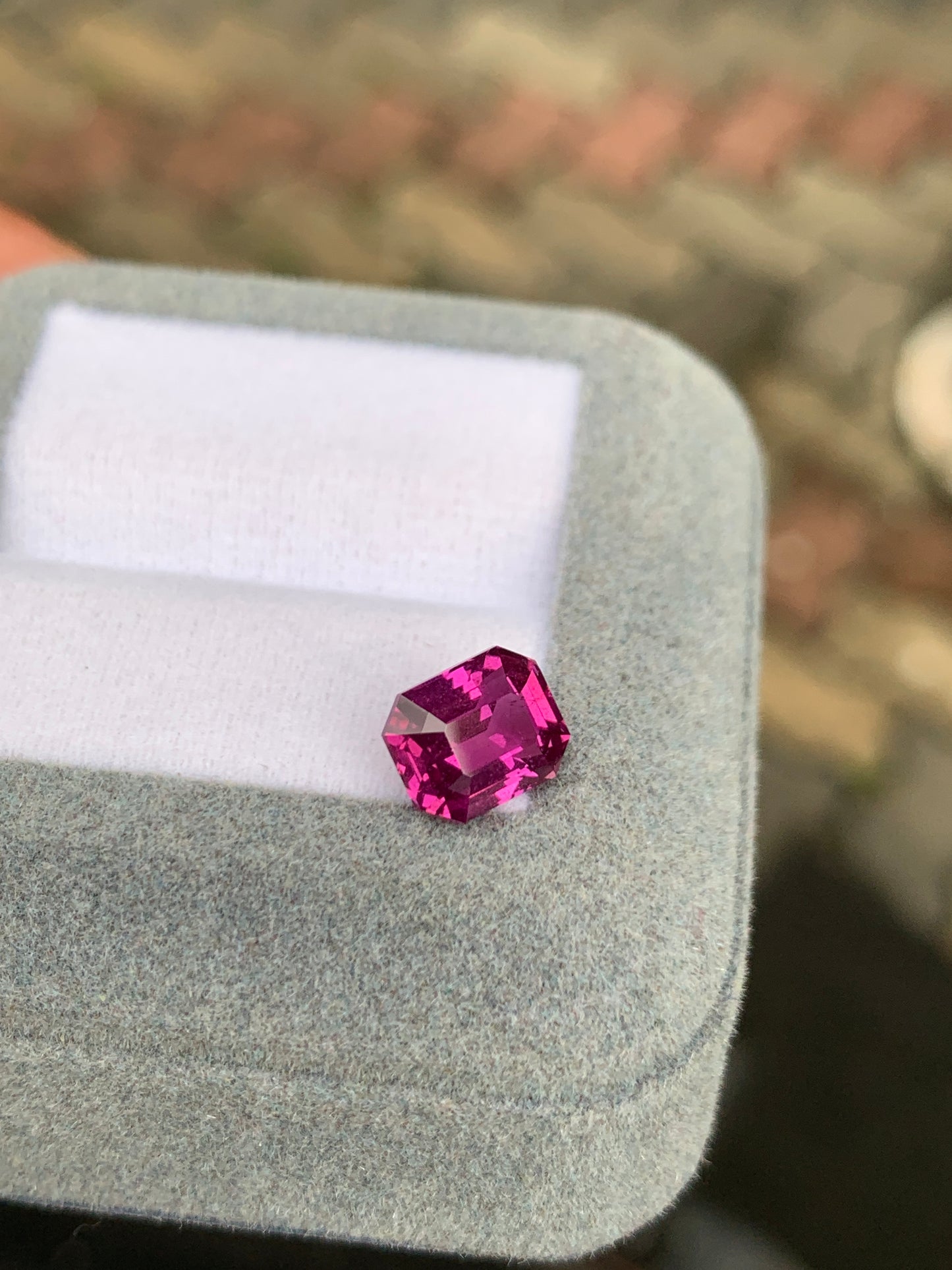 Un saphir rose vibrant de 3,01 ct avec une taille émeraude de Bonnot Paris est élégamment présenté sur une boîte à bijoux en velours gris clair, avec un fond flou aux tons terreux.