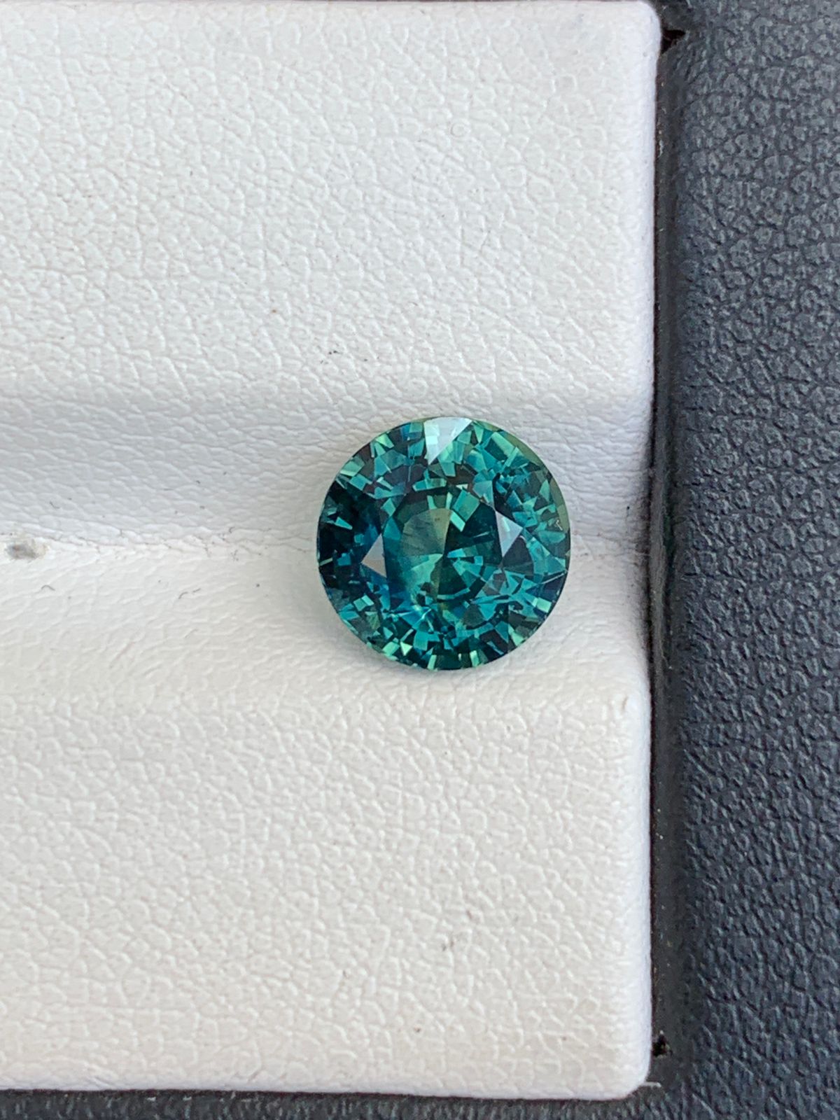 Le Saphir Teal • 3,6 carats de Bonnot Paris est une pierre précieuse ronde bleu sarcelle de 3,6 carats. Originaire de Madagascar, il éblouit par ses facettes complexes sur un fond texturé blanc et gris foncé, créant des reflets lumineux fascinants.
