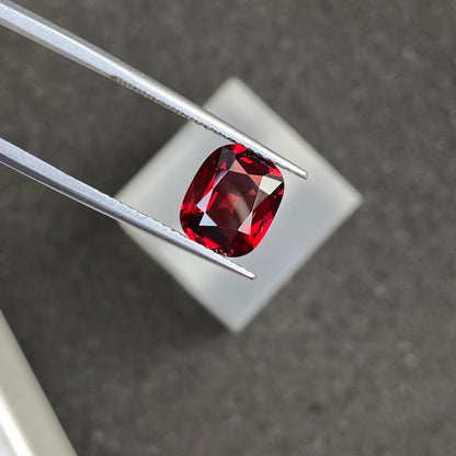 Une paire de pinces maintient le Spinelle Rouge · 2,3 ct de Bonnot Paris sur un fond gris flou, mettant en valeur la clarté de la pierre précieuse taille coussin et sa couleur rouge foncé sous un éclairage focalisé.