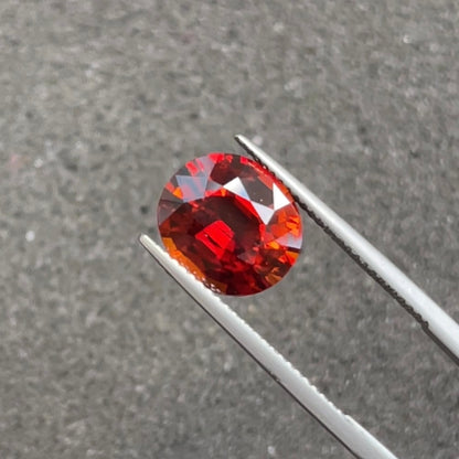 Une pierre précieuse ronde et polie Bonnot Paris Grenat Spessartite · 6,15 ct est serrée dans une pince métallique sur un fond gris texturé, ses facettes rouge foncé reflétant la lumière pour mettre en valeur sa couleur vibrante et sa clarté.