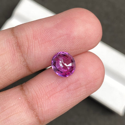 Une main tient le « Saphir Rose · 2,03 ct » de Bonnot Paris, une petite pierre violette ronde et facettée qui scintille sous la lumière. L'arrière-plan est flou et grisâtre.