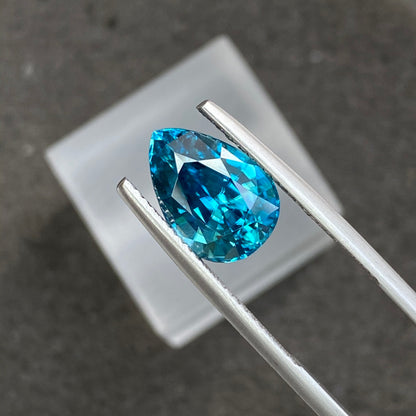 Une pierre précieuse bleue en forme de poire, le Zircon Bleu · 6,72 carats de Bonnot Paris, est maintenue entre des pinces en argent sur un fond sombre, avec un carré blanc flou en arrière-plan. La pierre scintille, révélant des facettes qui reflètent la lumière.