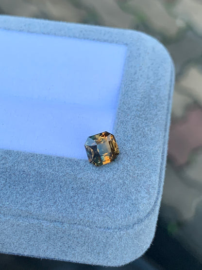 Un Saphir bicolore de 1,60 ct de [NOUVEAU] Bonnot Paris, aux tons ambrés et multifacettes, repose avec une lueur orange chaleureuse sur le coin d'une boîte à bijoux grise à la texture douce sur un fond flou.