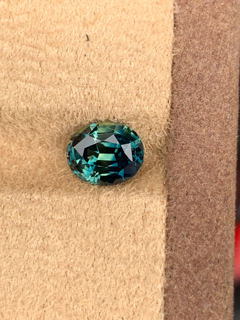 La pierre précieuse Saphir Teal de Bonnot Paris, de 2,7 carats, présente un design rond et facetté sur une surface beige texturée et une couleur vibrante et profonde. Ses tailles complexes reflètent brillamment la lumière, créant un éclat enchanteur.