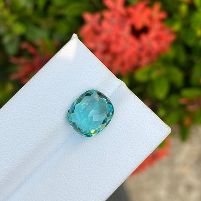 La Tourmaline Paraïba · 4,08ct de Bonnot Paris, une pierre précieuse bleue taille coussin, est présentée sur une surface blanche avec un fond flou de feuillage vert et de fleurs rouges.