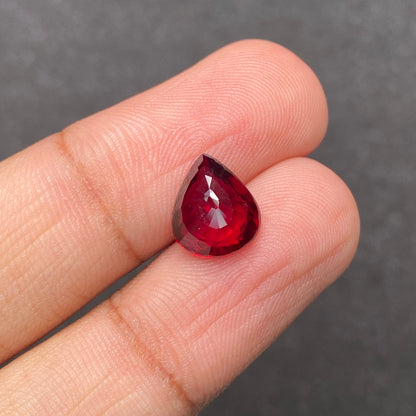Une main tient le Rubis du Mozambique · 4,03 ct de Bonnot Paris, une petite pierre précieuse rouge en forme de larme avec une surface facettée, sur un fond gris texturé.