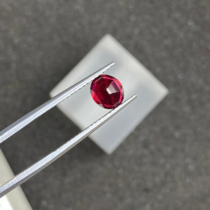 Une paire de pinces métalliques maintient la pierre précieuse Spinelle Rouge · 1,25 ct de Bonnot Paris, une pierre précieuse rouge foncé de forme ovale, sur un fond gris flou avec un carré blanc en dessous.