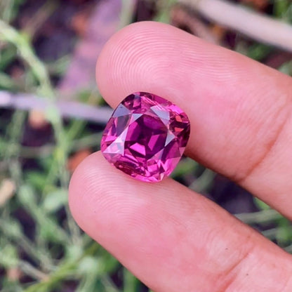 Une main tient la Tourmaline Rose, une pierre précieuse taille coussin de 6,21 carats de Bonnot Paris, ses facettes rose foncé scintillant sur un fond herbeux.