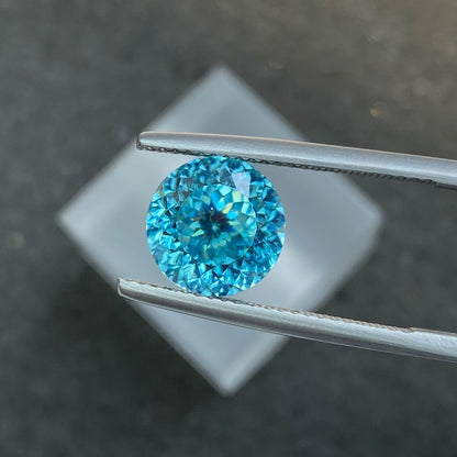 Une pince en argent maintient le Zircon Bleu · 7,56ct de Bonnot Paris sur un fond sombre flou, avec ses facettes bleues scintillant à la lumière.