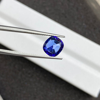Gros plan sur la tanzanite Bonnot Paris de 2,44 carats, une pierre précieuse bleue taille coussin, maintenue par une pince métallique sur un fond flou. Ses facettes reflètent la lumière, mettant en valeur sa teinte bleu profond.