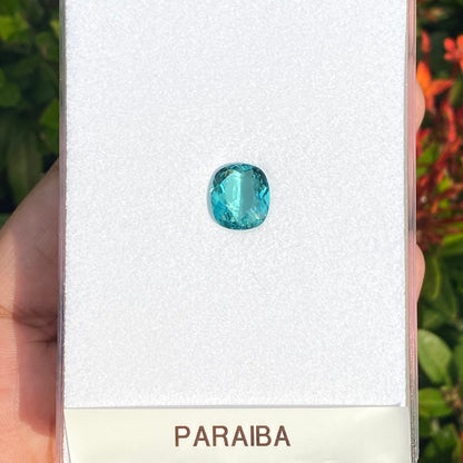 Quelqu'un tient une carte Bonnot Paris Tourmaline Paraïba · 4,08 ct comportant une petite pierre précieuse bleue transparente étiquetée « PARAIBA » devant un feuillage vert avec des fleurs orange.