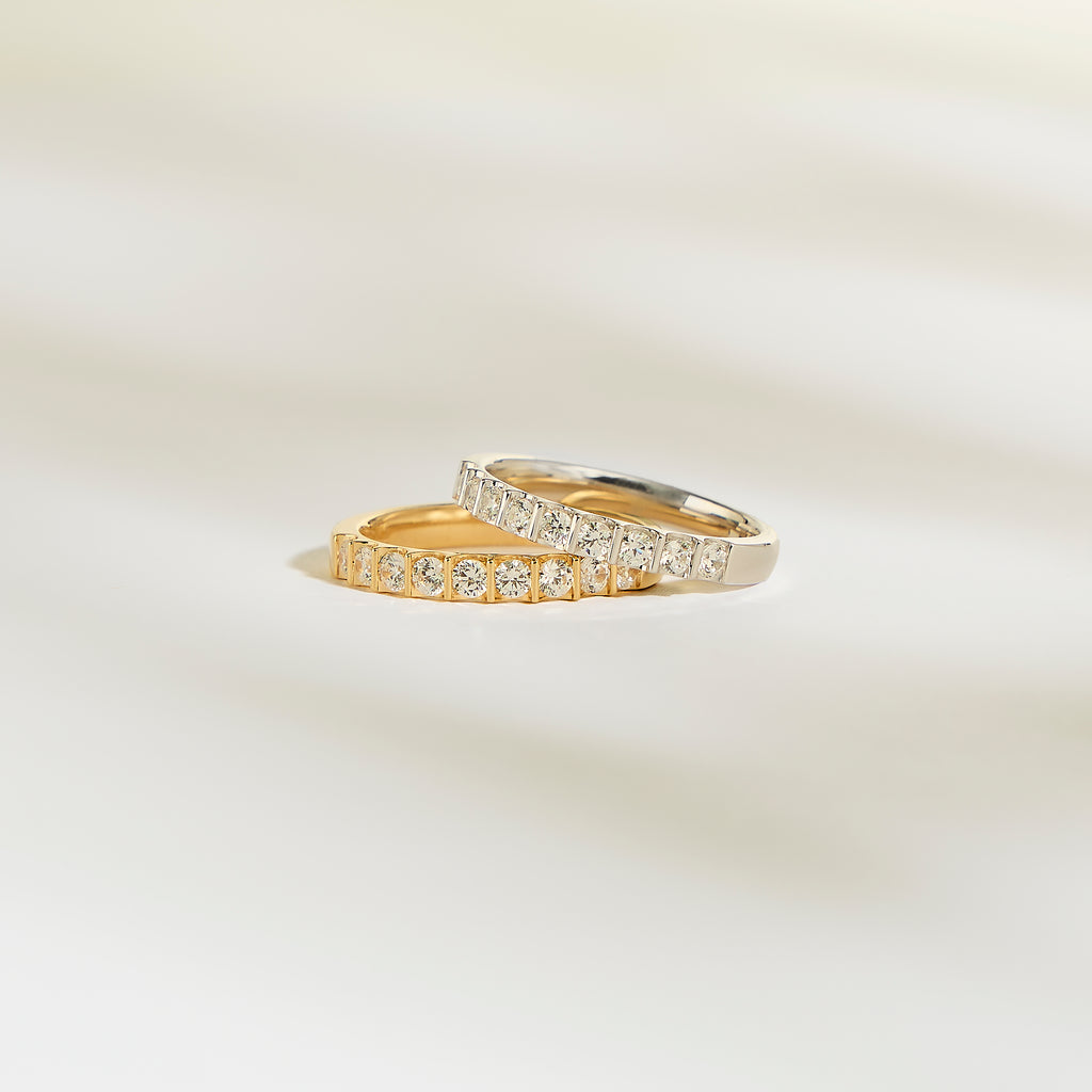 Les bagues Alliance La Majestueuse de Bonnot Paris, l'une en or et l'autre en argent, arborent des diamants brillants sur une surface douce et claire. Le fond légèrement flouté rehausse le luxe serein de la couleur clarté G/H SI.