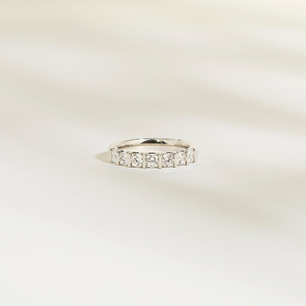 L'Alliance La Subtile de Bonnot Paris est une bague en argent sertie de pierres taille carrée, semblables à des diamants. Son éclat brillant évoque un luxe discret, issu d'une collection exquise. Elle repose sur une surface lisse et légère, avec des ombres douces et floues en arrière-plan.