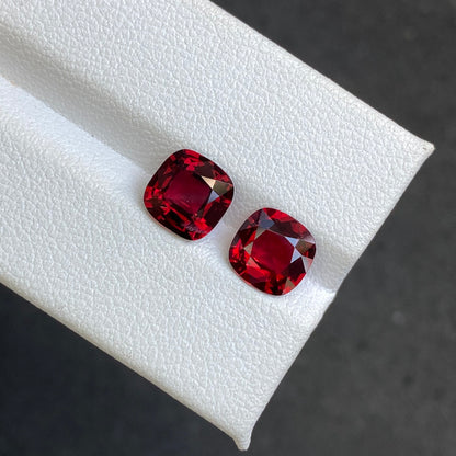 Deux pierres précieuses Bonnot Paris Spinelle Rouge de 3,49 carats, taille coussin, sont mises en valeur sur une surface blanche texturée, d'un rouge profond et vibrant. Elles se juxtaposent sur un fond gris foncé, accentuant leur éclat.