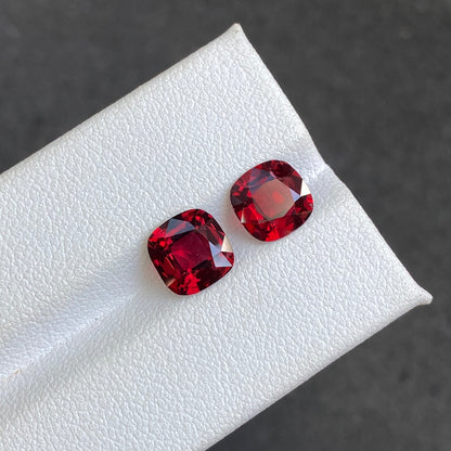 Deux spinelles rouges (3,49 carats) de Bonnot Paris, taillées en carré et dotées d'une finition brillante d'un rouge profond, reposent sur une surface blanche texturée. Un fond gris foncé flou rehausse leur teinte vive.
