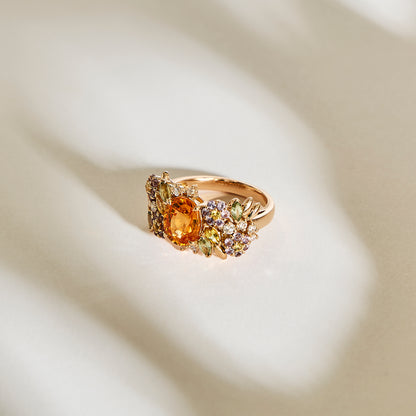 La bague Golden Bloom - Grenat Mandarin de Bonnot Paris présente une spessartite grenat centrale entourée d'une fleur de couleur de pierres violettes et vertes plus petites dans un design floral exquis, le tout reposant sur une surface texturée doucement éclairée.