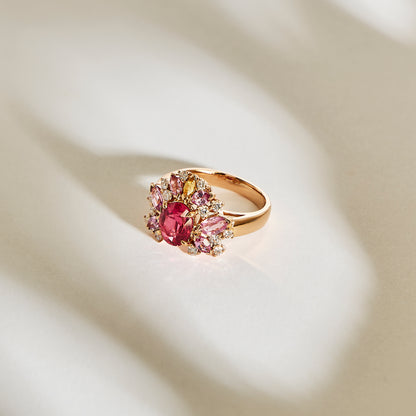 La bague en or Bonnot Paris "Rose Bloom - Spinelle Mahenge" présente une spinelle Mahenge centrale proéminente, entourée de pierres roses et claires plus petites, reposant élégamment sur une surface doucement éclairée et légère.
