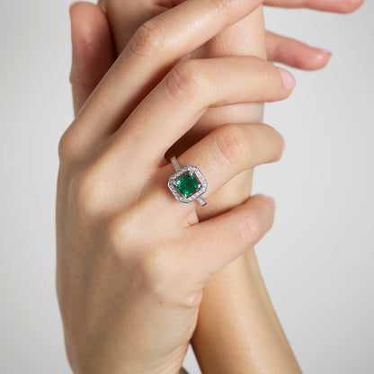 Gros plan sur une main portant la Bague Émeraude • 1,22 carat de Bonnot Paris. Sa pierre zambienne taille émeraude est entourée d'un halo de diamants sertis dans un élégant métal blanc, exprimant un style Art déco. La main au poli neutre en saisit une autre sur un fond sobre.