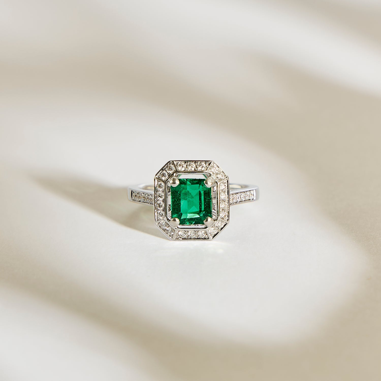La Bague Emeraude • 1,22ct de Bonnot Paris présente une pierre précieuse verte taille émeraude évoquant le style Art Déco, entourée d'un halo de diamants étincelants sur une surface argentée aux ombres douces.