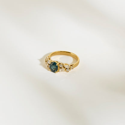 La Bague Saphir Teal de Bonnot Paris • 1,14 carat est ornée d'un saphir bleu sarcelle central entouré de petits diamants blancs. Sur un fond crème clair, ses détails raffinés et son design élégant en font une bague de fiançailles idéale.