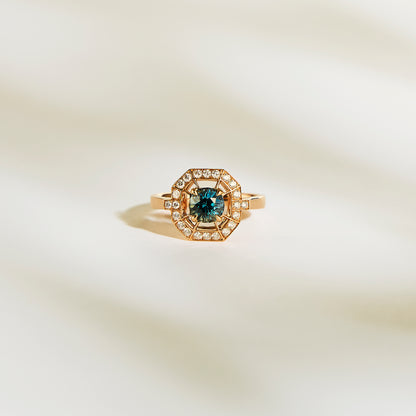 La Bague Saphir Teal de Bonnot Paris • 1,16 ct est une superbe bague de fiançailles en or avec un saphir sarcelle central et un halo de diamants blancs étincelants, présentés sur une surface douce et claire.