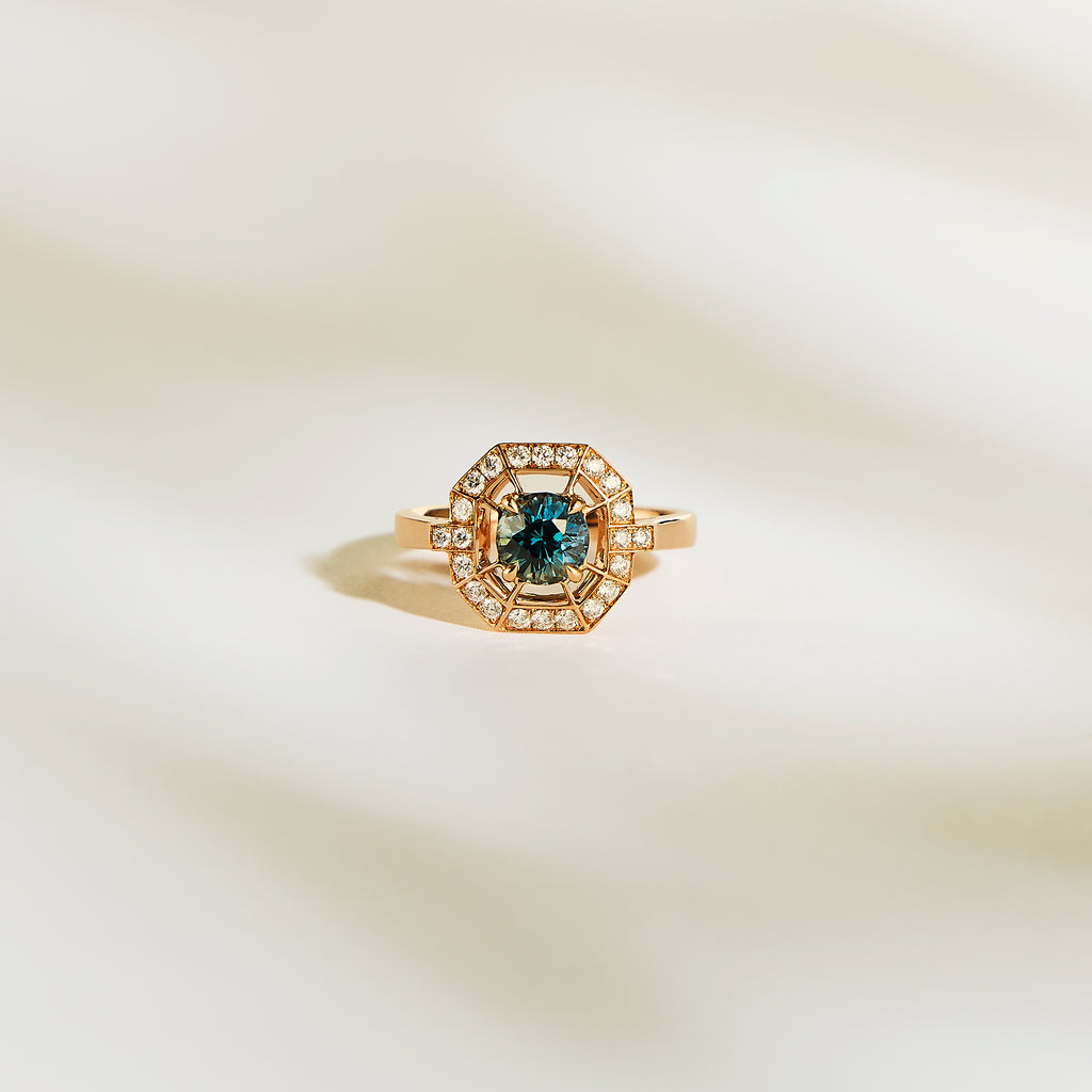 La Bague Saphir Teal de Bonnot Paris • 1,16 ct est une superbe bague de fiançailles en or avec un saphir sarcelle central et un halo de diamants blancs étincelants, présentés sur une surface douce et claire.
