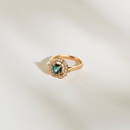 La Bague Saphir Teal • 1,16ct de Bonnot Paris présente un large saphir bleu sarcelle entouré de diamants blancs étincelants, élégamment présenté sur un fond doux et clair.
