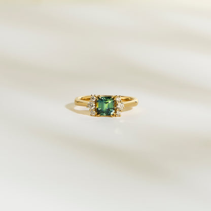 La Bague Bonnot Paris Saphir Teal • 1,24 ct présente une pierre précieuse sarcelle carrée flanquée de petits diamants blancs étincelants sur une bande lisse et de couleur claire.