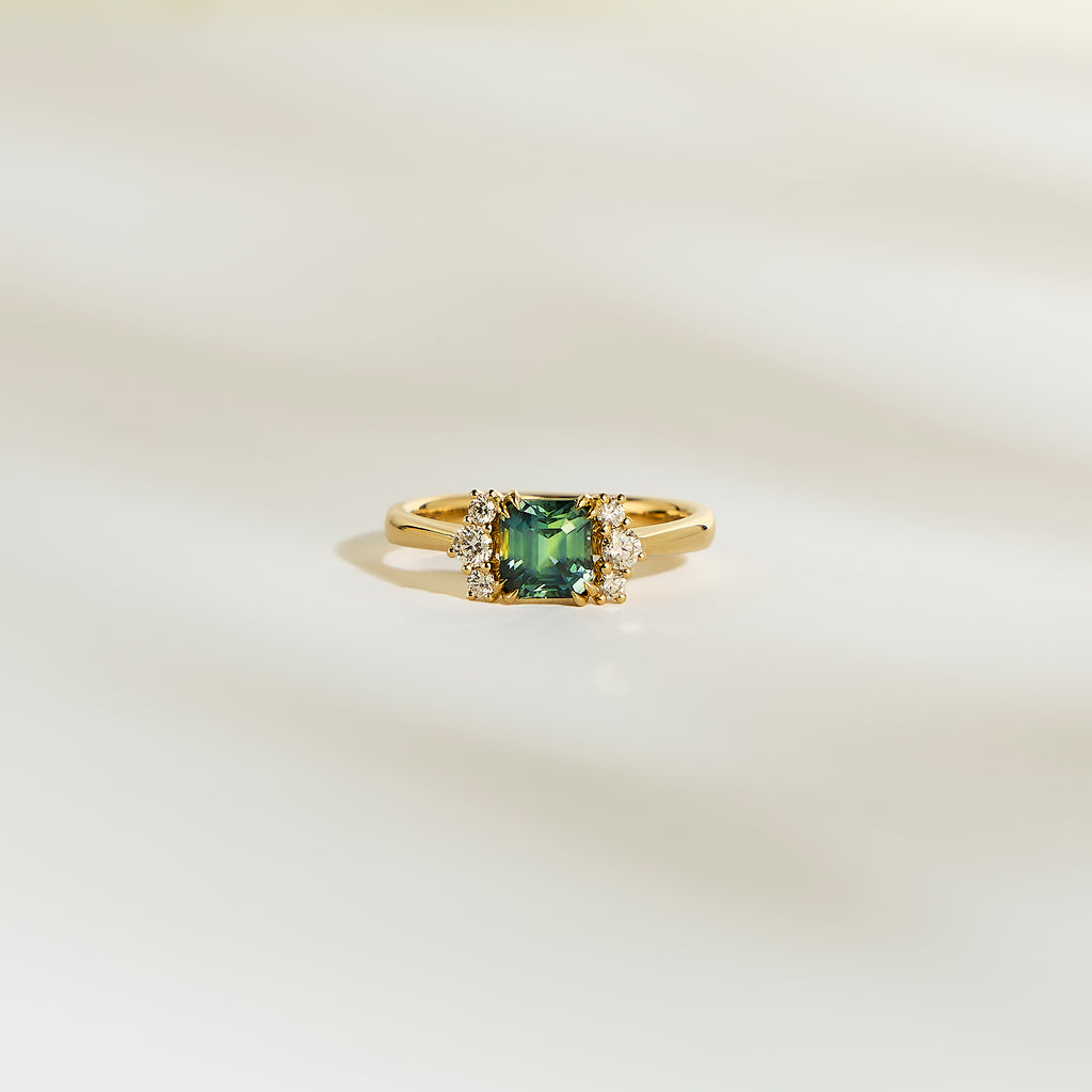 La Bague Bonnot Paris Saphir Teal • 1,24 ct présente une pierre précieuse sarcelle carrée flanquée de petits diamants blancs étincelants sur une bande lisse et de couleur claire.