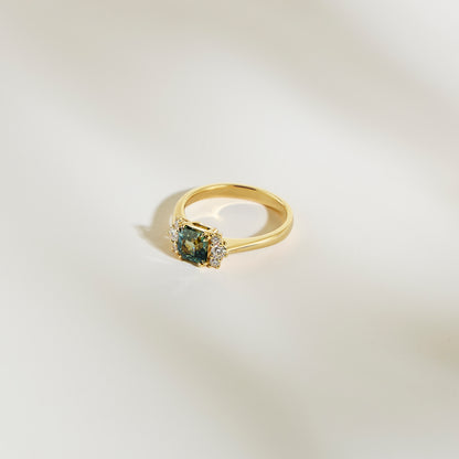 La Bague Saphir Teal • 1,24ct de Bonnot Paris, une bague en or avec un saphir sarcelle taille carrée en son centre et entouré de petits diamants blancs, est présentée sur une surface lisse de couleur claire.