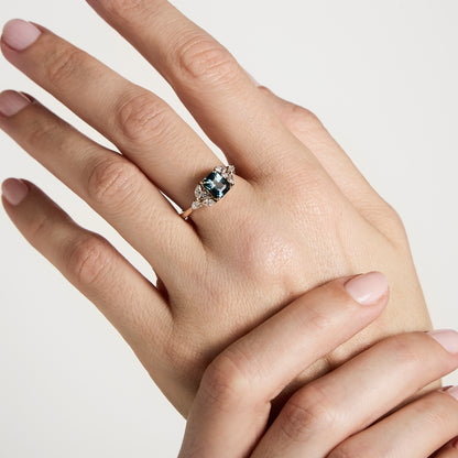 Une main aux ongles manucurés arbore une Bague Saphir Teal Bonnot Paris de 1,56 carat. La bague est ornée d'un grand saphir rectangulaire turquoise encerclé de petits diamants blancs. Les doigts, élégamment posés, se détachent sur un fond blanc uni, mettant en valeur cette magnifique bague de fiançailles.