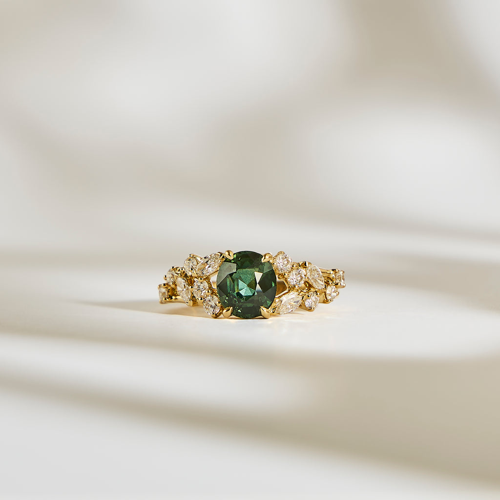 La Bague Saphir Teal • 2,07ct de Bonnot Paris, une bague en or avec un centre en saphir bleu sarcelle saisissant entouré de pierres claires, repose élégamment sur une surface crème doucement éclairée avec des ombres douces.